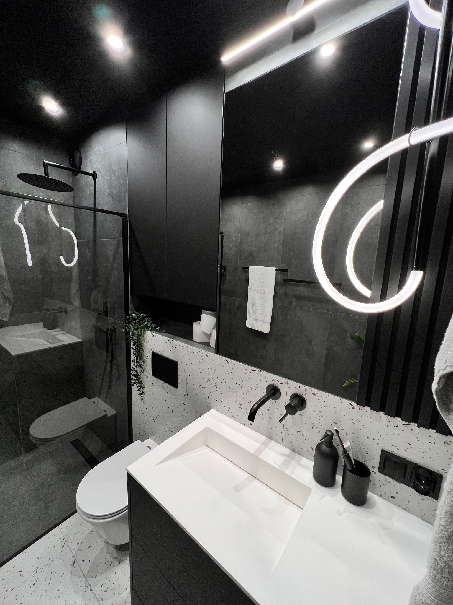 Nowoczesna łazienka w czerni i bieli: minimalistyczna umywalka, deszczownica za szklaną ścianką, designerskie oświetlenie i płytki lastryko.