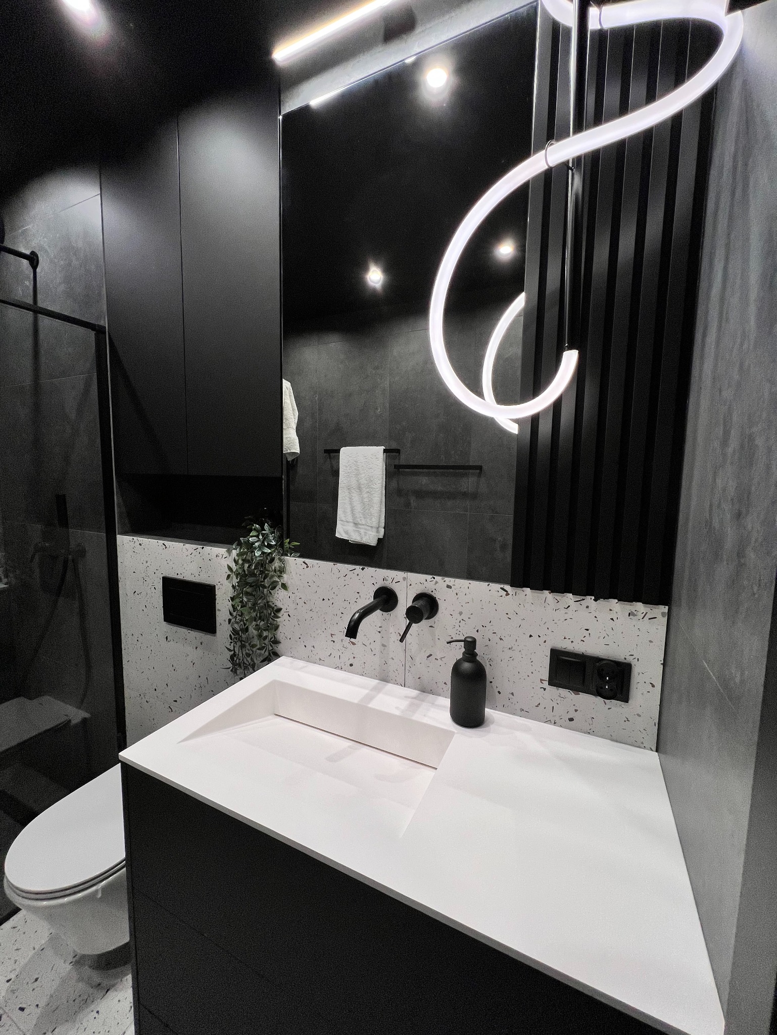 Nowoczesna łazienka w czerni i bieli: biała umywalka z czarną armaturą, lustro z dekoracyjnym oświetleniem LED, szare płytki i akcenty roślinne. Minimalistyczny design.