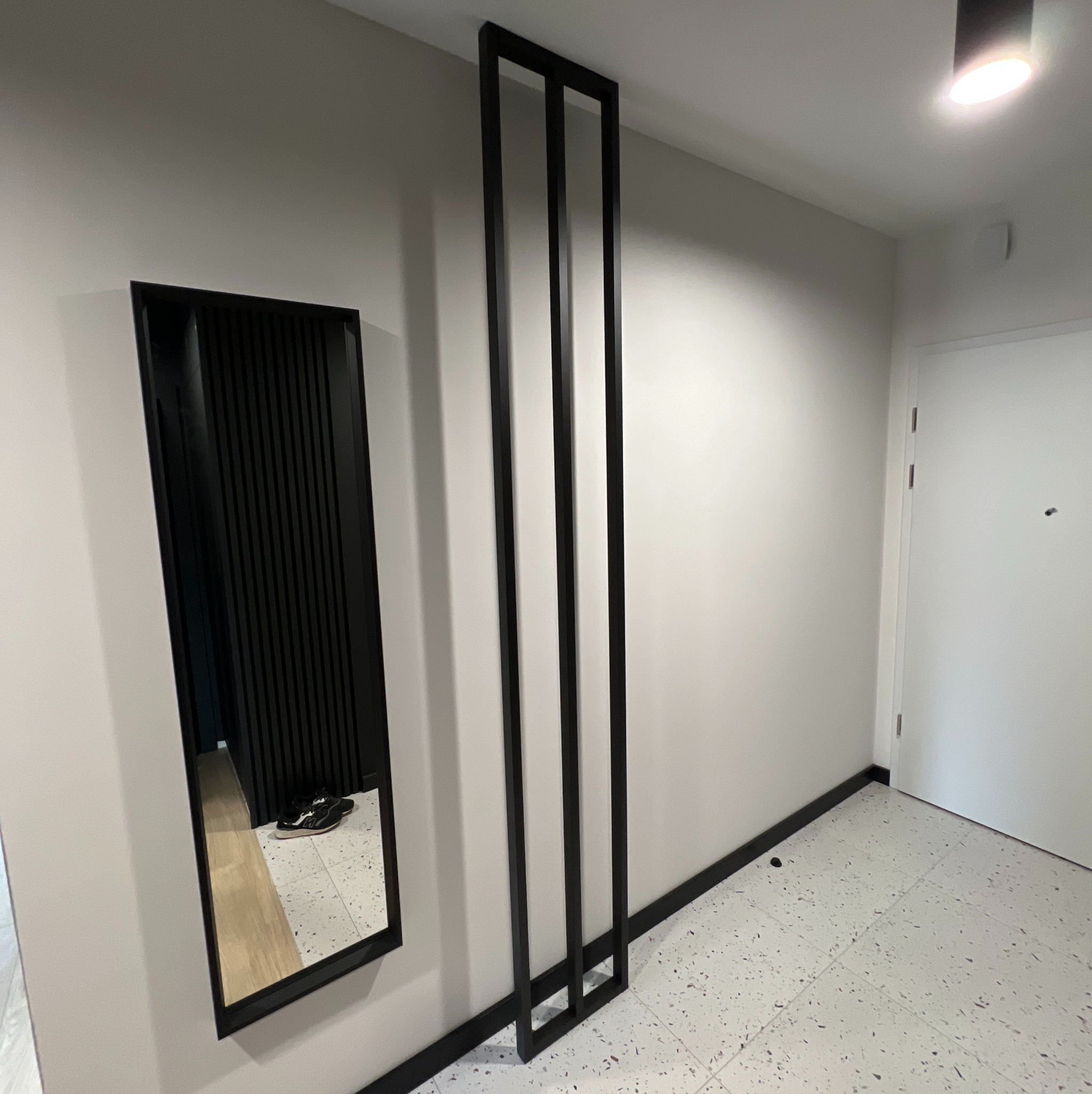 Nowoczesny przedpokój z lustrem w czarnej ramie i dekoracyjną ścianką z czarnych listew. Białe ściany i podłoga w stylu lastryko. Minimalistyczny design.