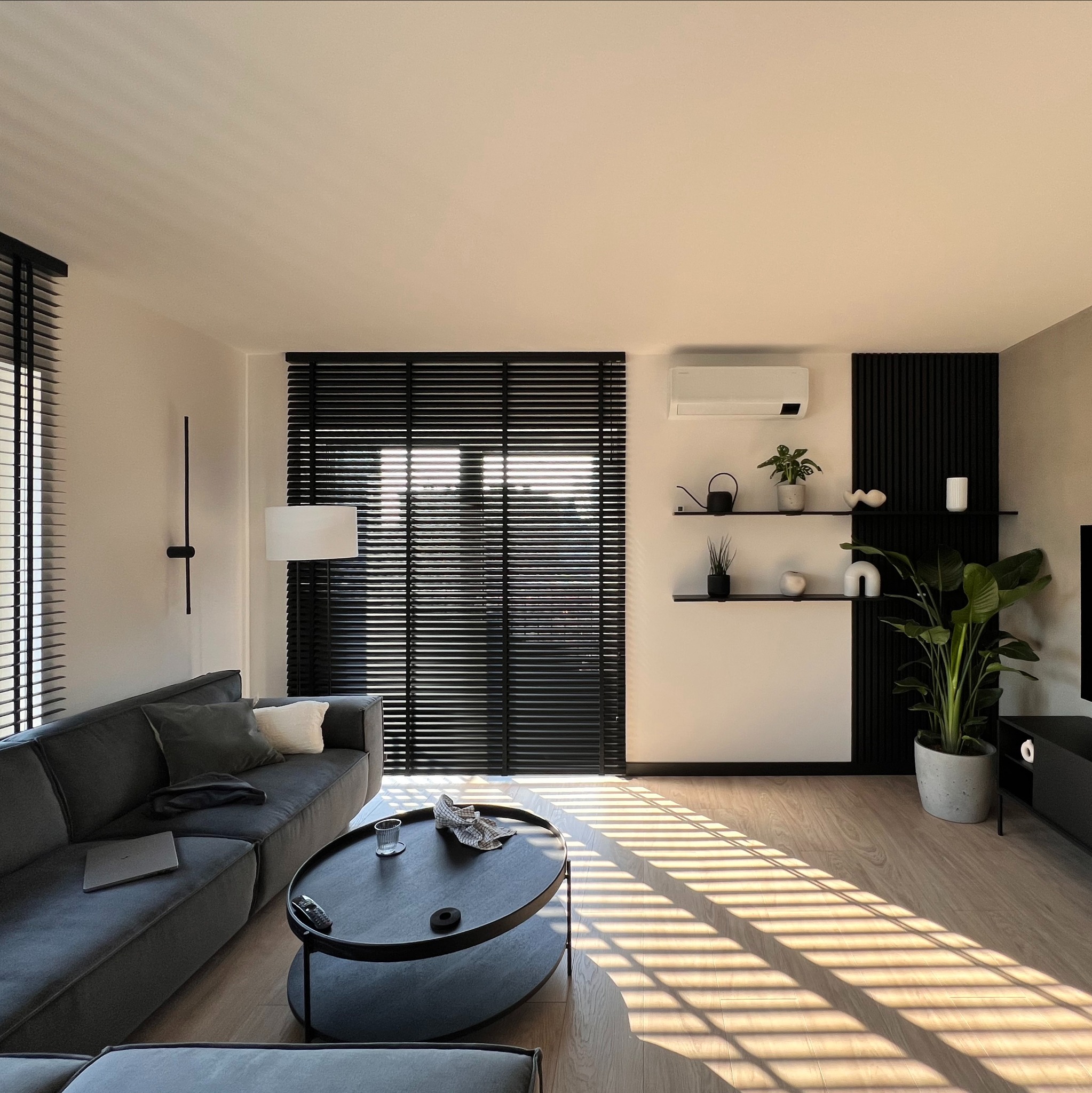 Nowoczesny salon z szarym narożnikiem, okrągłym stolikiem i drewnianymi żaluzjami, przez które wpada ciepłe światło słoneczne. Minimalistyczne półki z dekoracjami.