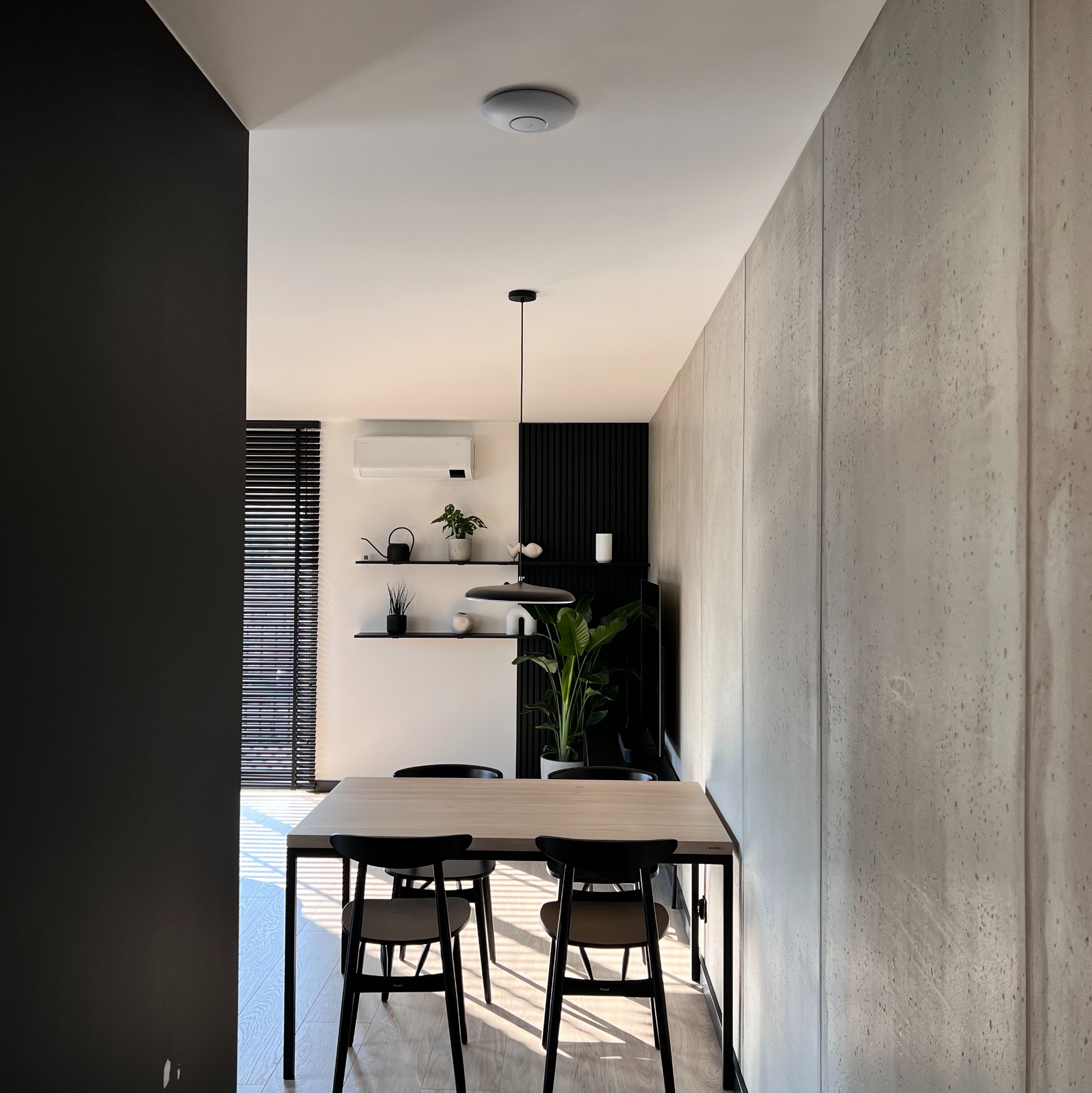 Nowoczesne wnętrze jadalni z betonową ścianą, drewnianym stołem i czarnymi krzesłami. Minimalistyczny design z akcentami roślinnymi i oświetleniem.