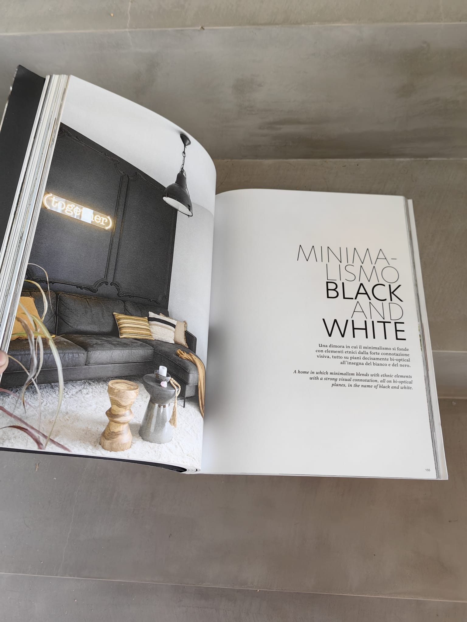Otwarta książka prezentuje wnętrze w stylu minimalistycznym: czarna ściana z dekoracyjną sztukaterią, szara sofa, dywan i neon 'together'.