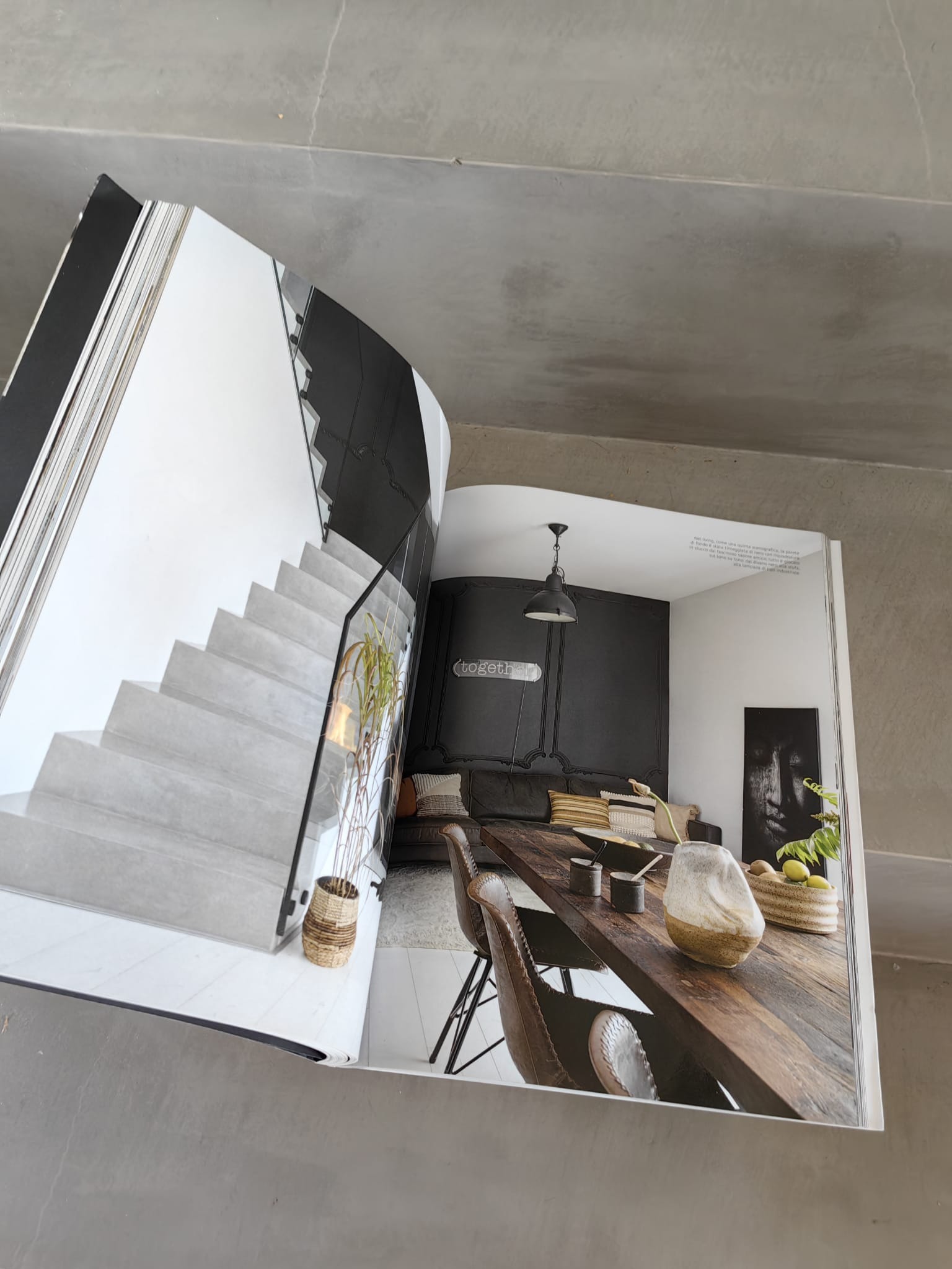 Otwarte wydanie magazynu wnętrzarskiego prezentuje nowoczesne schody z szarego betonu i minimalistyczną jadalnię z drewnianym stołem oraz czarną ścianą w tle.