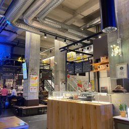 BUILDUPGROUP SPÓŁKA Z OGRANICZONĄ ODPOWIEDZIALNOŚCIĄ - Wnętrze hali targowej z widocznymi instalacjami wentylacyjnymi pod sufitem, betonowymi kolumnami z napisami, oświetleniem punktowym, ladą chłodniczą i neonowym szyldem.