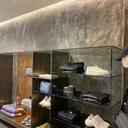 BUILDUPGROUP SPÓŁKA Z OGRANICZONĄ ODPOWIEDZIALNOŚCIĄ - Wnętrze sklepu odzieżowego z półkami ekspozycyjnymi, na których znajdują się ubrania i buty marki Givenchy, tło stanowi ściana o surowej, betonowej fakturze, oświetlenie punktowe. Manekin w czapce...