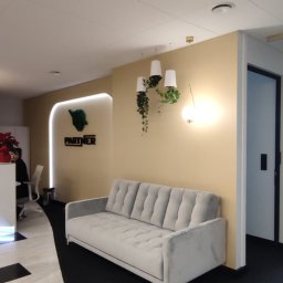 BUILDUPGROUP SPÓŁKA Z OGRANICZONĄ ODPOWIEDZIALNOŚCIĄ - Jasnoszary welurowy sofa w recepcji biura, z wiszącymi roślinami w białych doniczkach i okrągłą lampą, na tle ściany w odcieniach beżu i logo firmy.