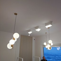 BUILDUPGROUP SPÓŁKA Z OGRANICZONĄ ODPOWIEDZIALNOŚCIĄ - Wnętrze w trakcie prac wykończeniowych z widocznymi dwoma wiszącymi, nowoczesnymi lampami kulistymi, zabezpieczonymi lampami sufitowymi, oraz pracownikiem w kombinezonie na tle okna z widokiem...