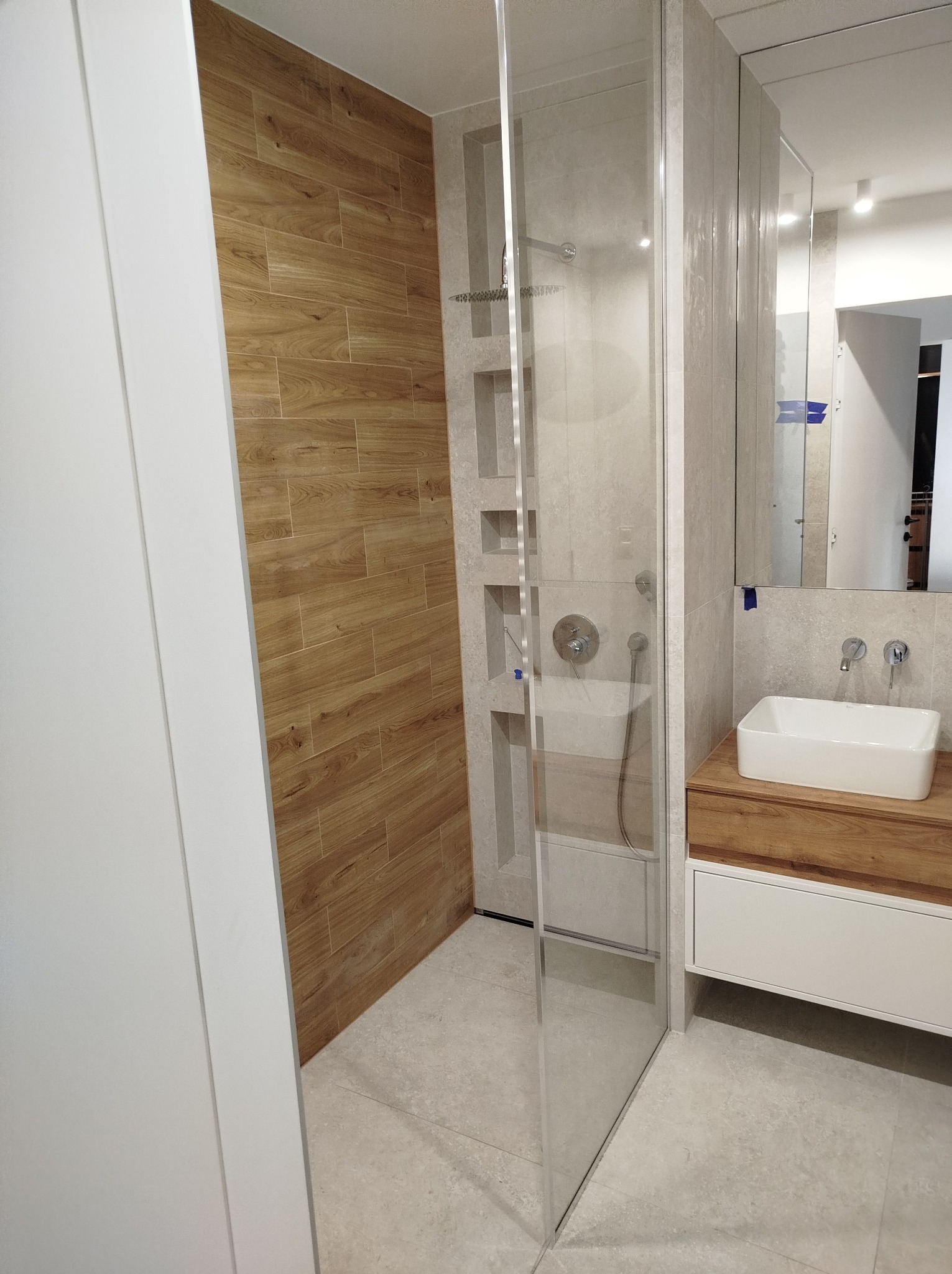 Nowoczesna łazienka z kabiną prysznicową typu walk-in, umywalką nablatową na drewnianej szafce i ścianą wyłożoną płytkami imitującymi drewno.