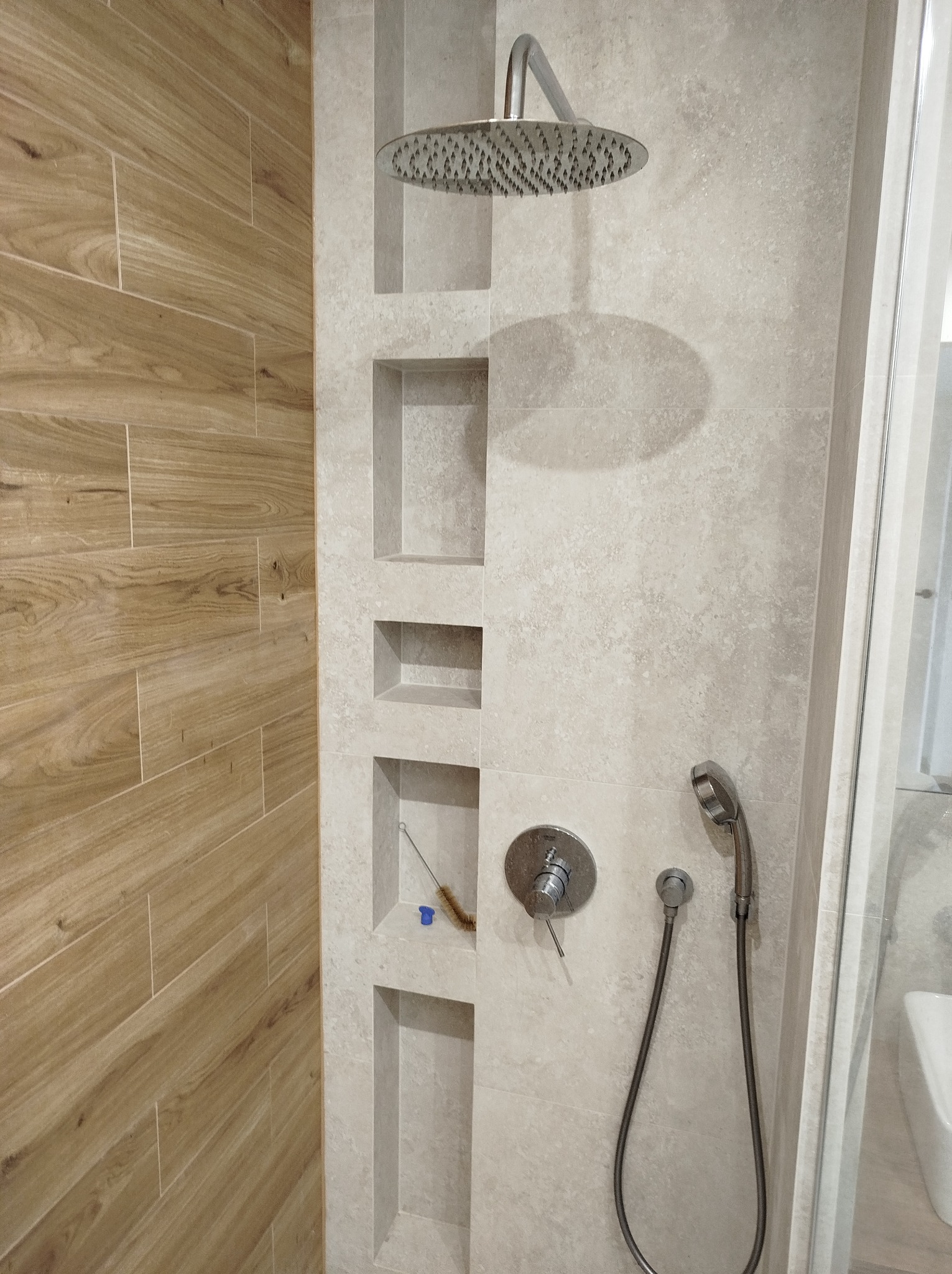 Nowoczesna kabina prysznicowa z drewnopodobnymi płytkami na ścianie, wnękami w ścianie wyłożonej jasnymi płytkami oraz deszczownicą i słuchawką prysznicową.