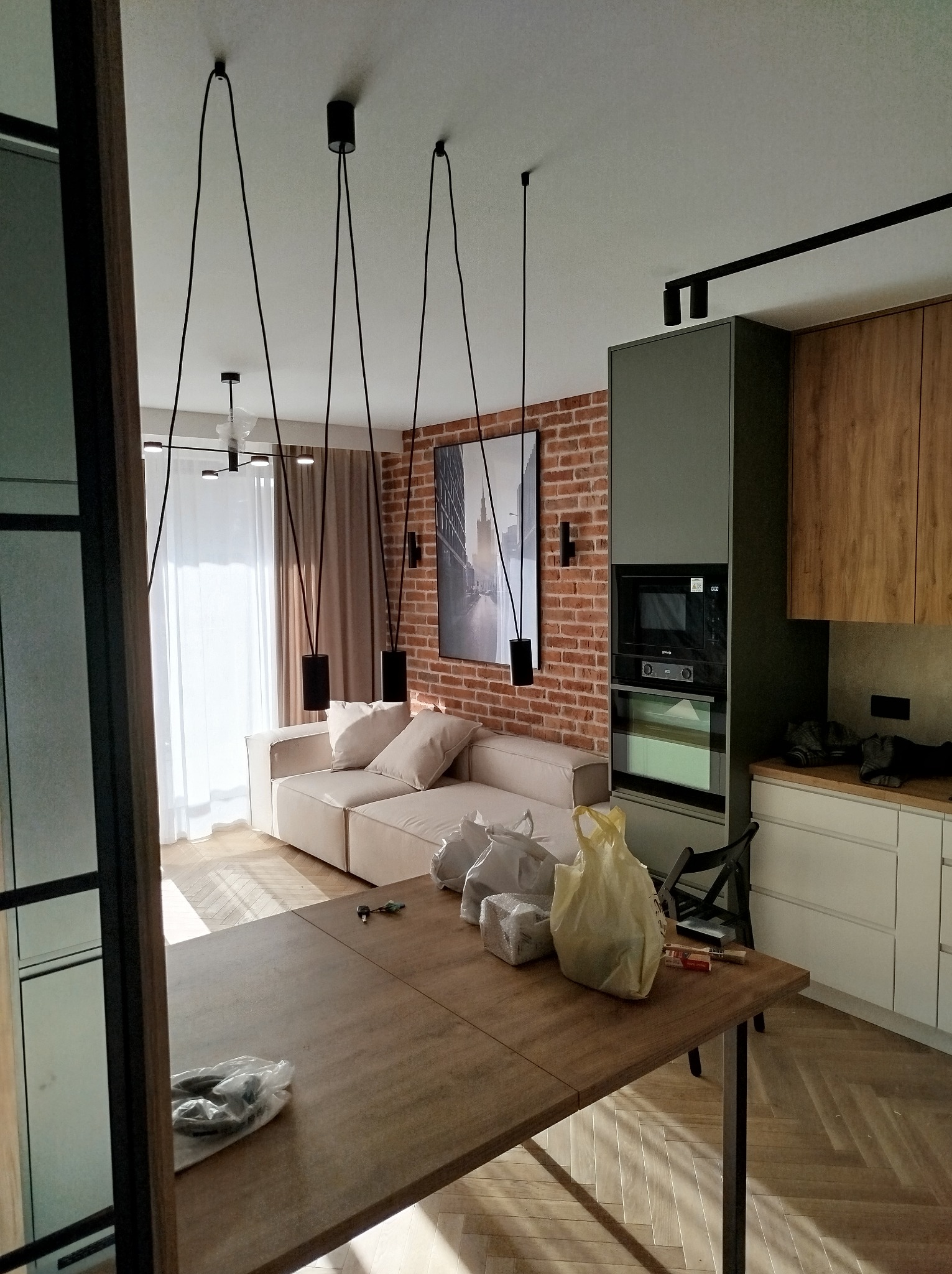 Wnętrze mieszkania w trakcie wykańczania: widoczny salon z sofą, lampami wiszącymi na długich kablach, ceglana ściana, zabudowa kuchenna oraz stół z materiałami budowlanymi i narzędziami.