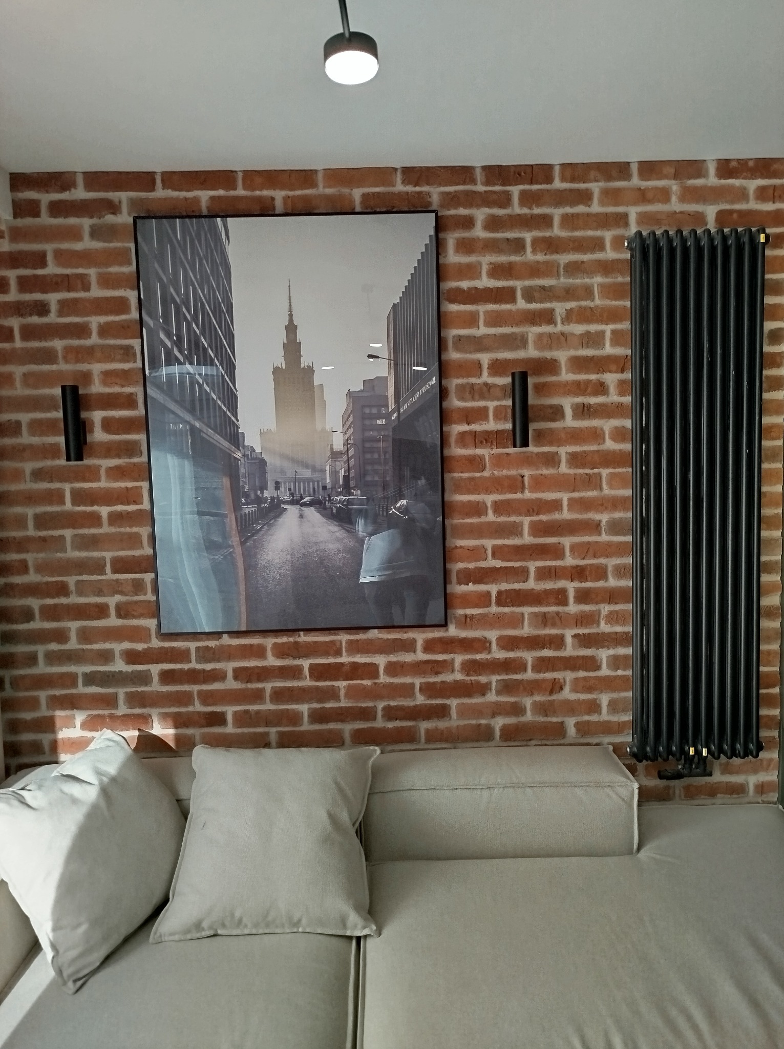 Ceglana ściana z czarnym grzejnikiem żeberkowym i lampami ściennymi, nad jasną sofą z poduszkami, ozdobiona czarno-białym plakatem z motywem miejskim.