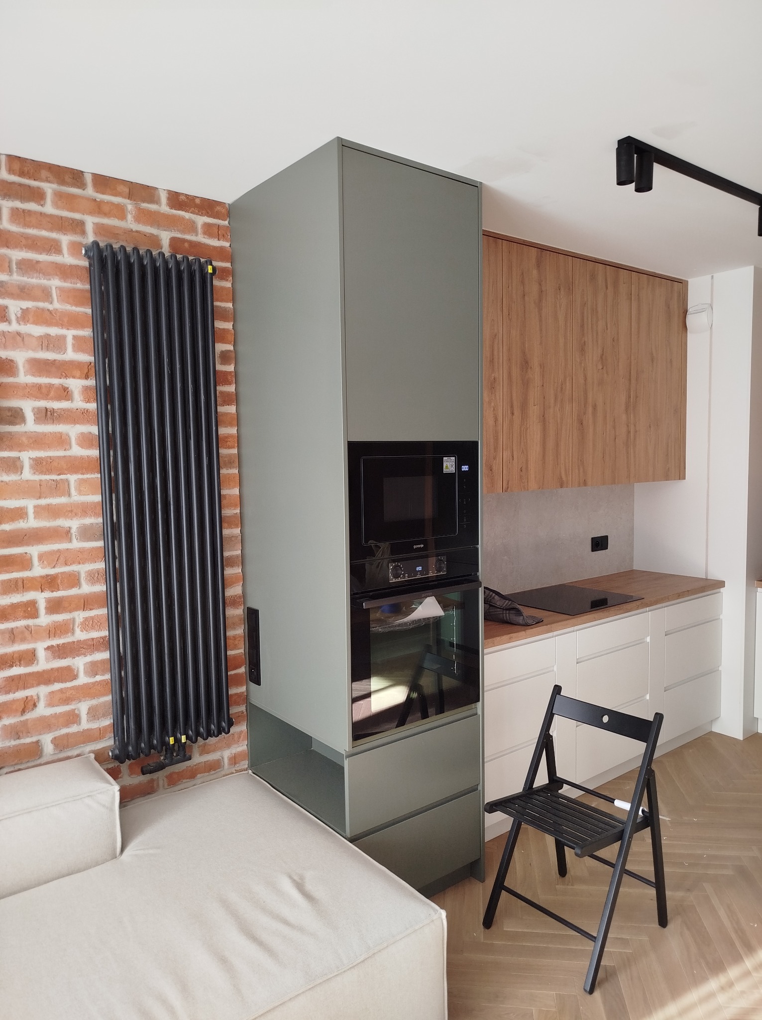 Nowoczesna kuchnia z zabudową w odcieniach szarości i drewna, widoczny piekarnik i mikrofalówka Gorenje, czarny grzejnik żeberkowy na tle ceglanej ściany, jasna sofa i czarne składane krzesło.
