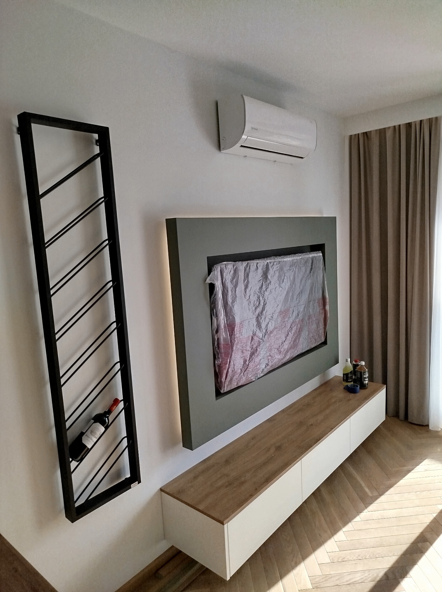 Nowoczesne wnętrze salonu z wiszącą szafką RTV, obudowanym telewizorem zabezpieczonym folią, klimatyzatorem i metalowym regałem na wino z jedną butelką, parkiet w jodełkę.