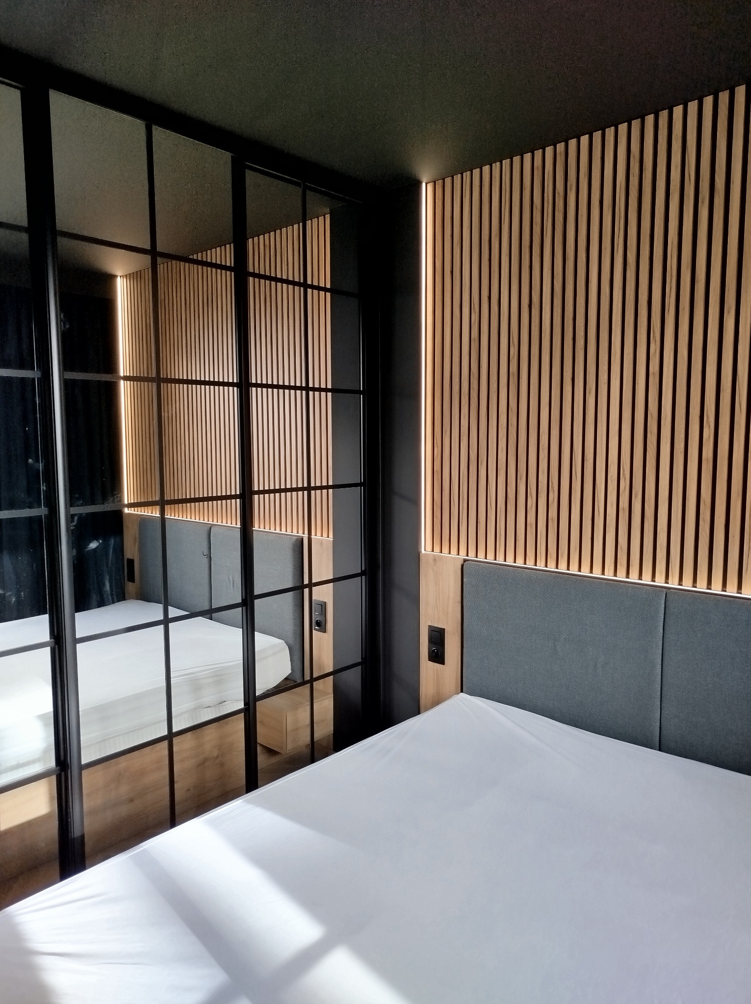 Nowoczesna sypialnia z drewnianą ścianą z lameli podświetlonych od tyłu, czarnym sufitem i ścianami, oraz łóżkiem z szarą tapicerką wezgłowia, widok przez przeszkloną ścianę z czarnymi szprosami.