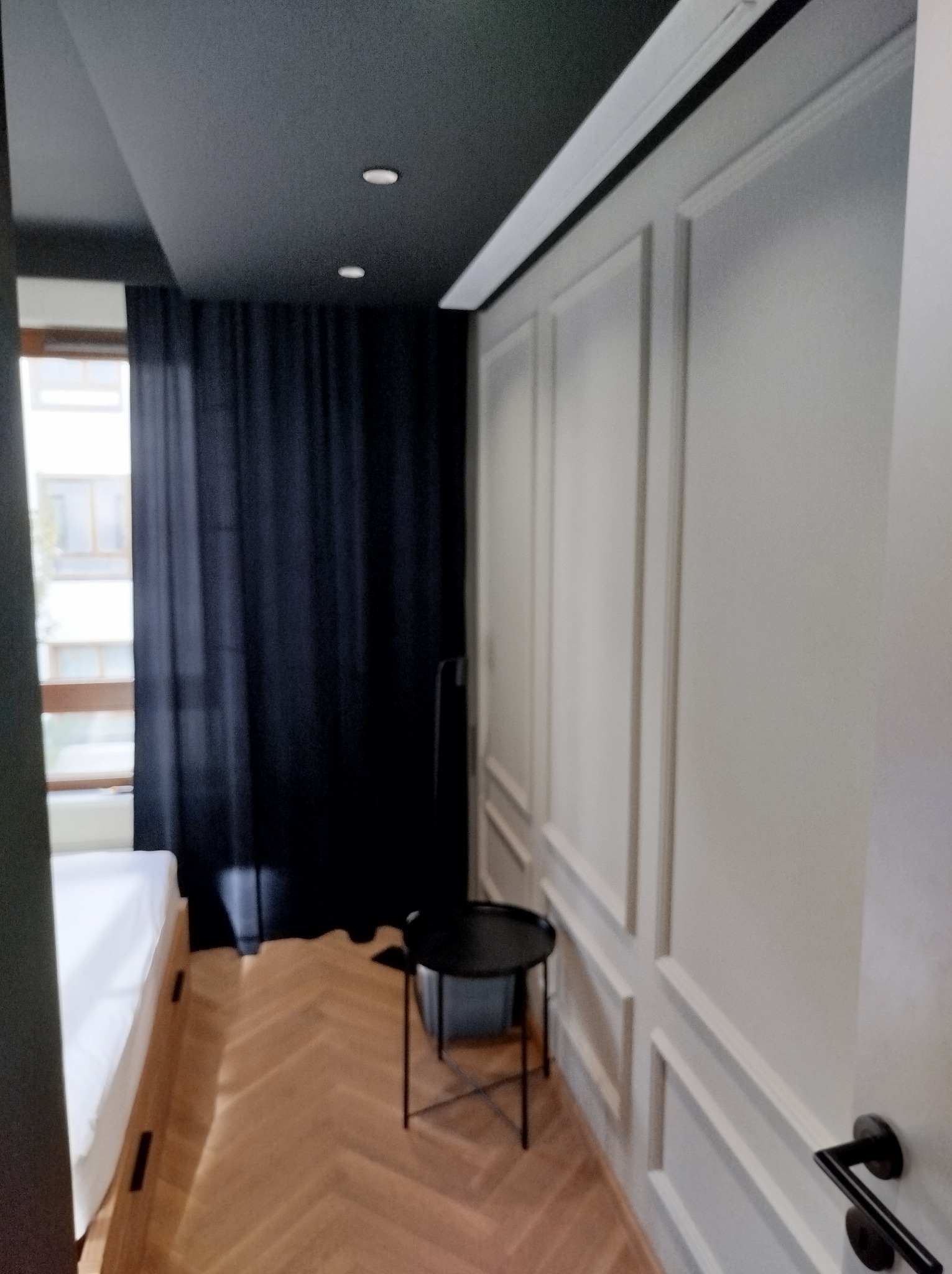 Eleganckie wnętrze sypialni z ciemnym sufitem, punktowym oświetleniem, parkietem w jodełkę, białą ścianą z dekoracyjnymi panelami i czarnymi zasłonami zaciemniającymi okno.