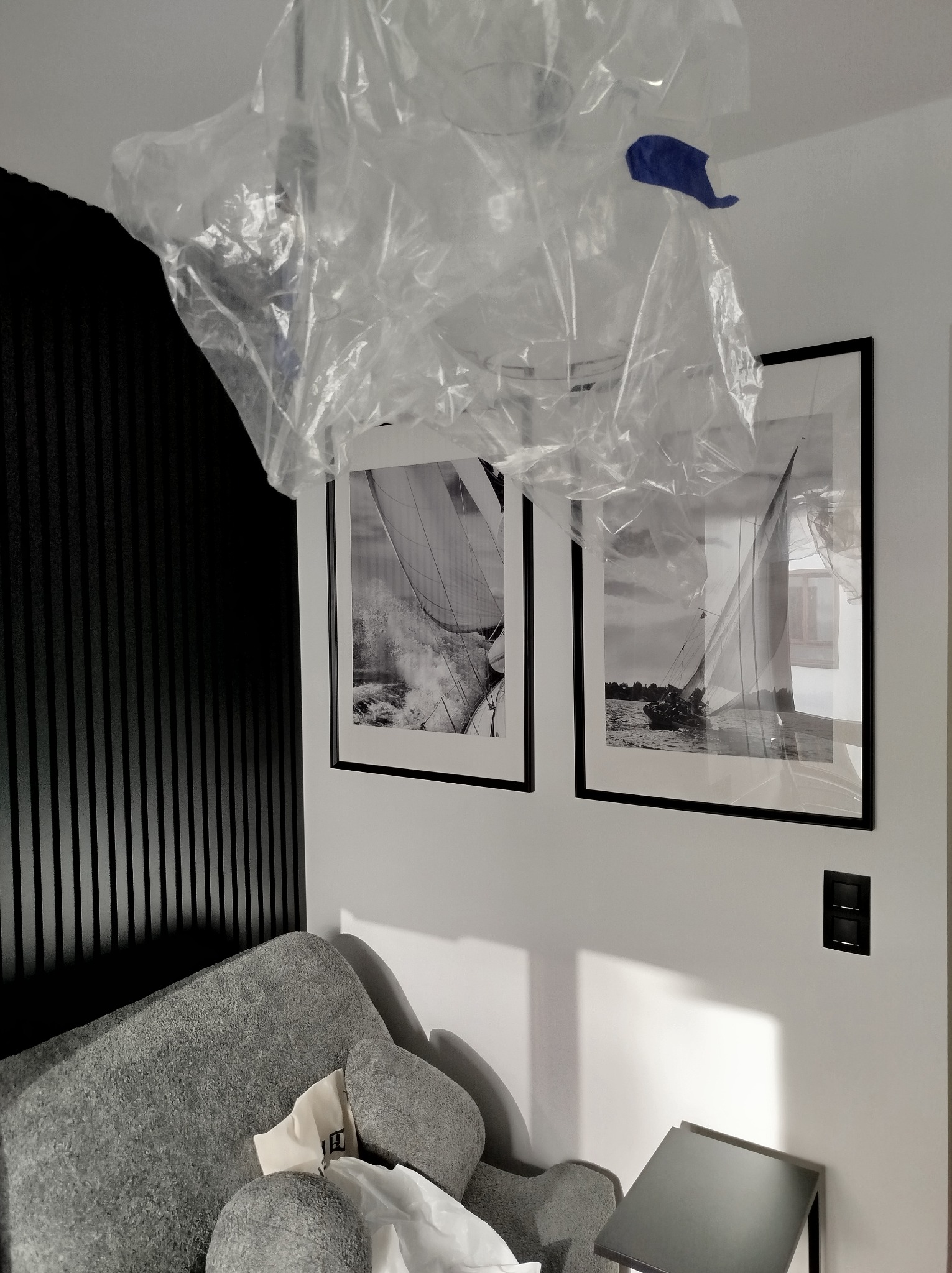 Ochrona lampy sufitowej folią malarską podczas prac remontowych, widoczne obrazy z motywem żaglówek na ścianie i fragment sofy.