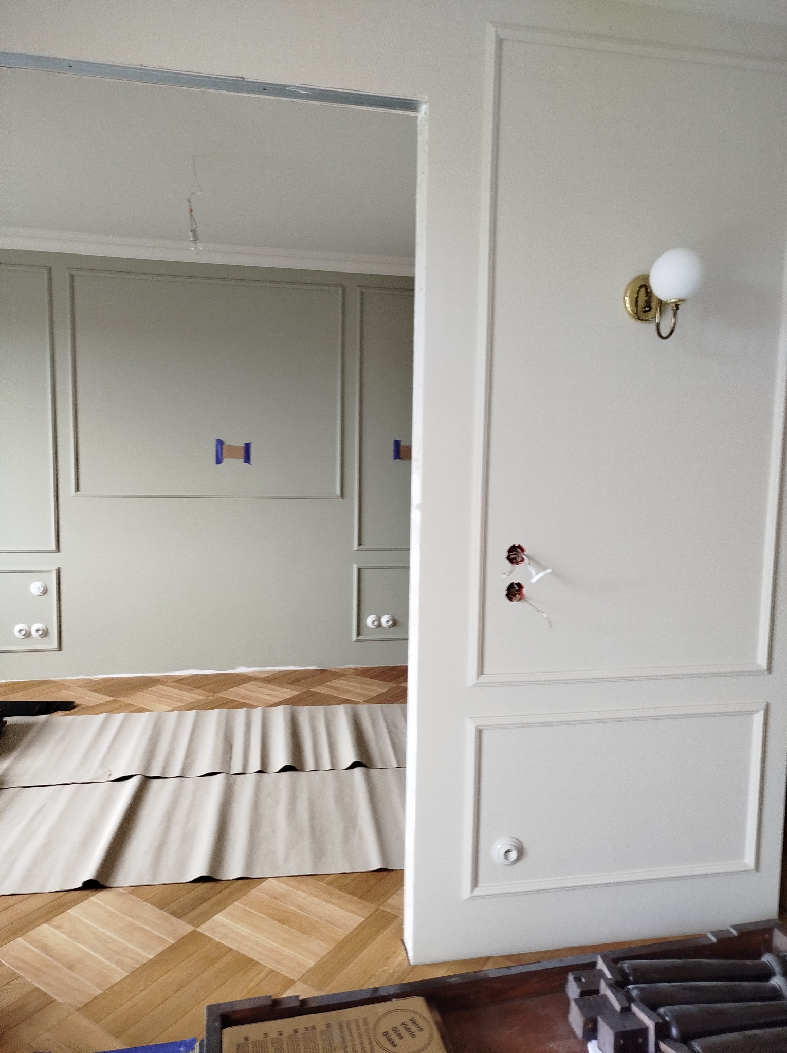 Pokój w trakcie prac wykończeniowych: zabezpieczona podłoga parkietowa, ściany z dekoracyjnymi panelami, widoczne gniazdka elektryczne, przewody do oświetlenia na ścianie i suficie, drzwi...