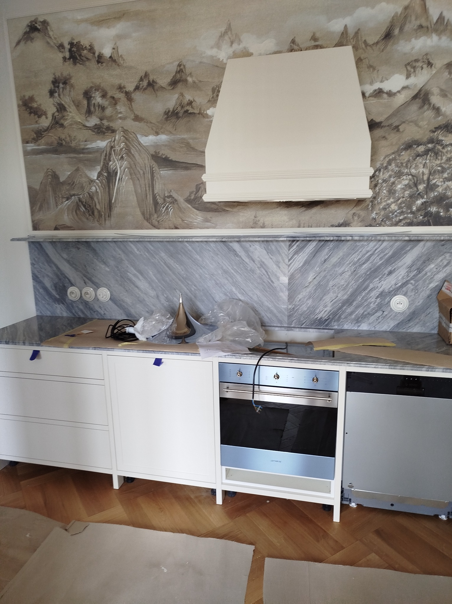 Montaż nowej kuchni z marmurowym blatem i piekarnikiem w zabudowie, widoczne zabezpieczenia blatu i podłogi kartonem, ściana z malowidłem krajobrazu.