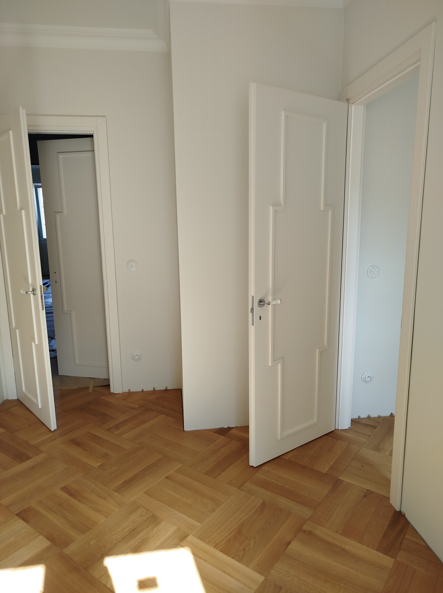 Jasne wnętrze z parkietem w jodełkę, otwarte białe drzwi z dekoracyjnymi panelami i widoczne gniazdka elektryczne na ścianie.