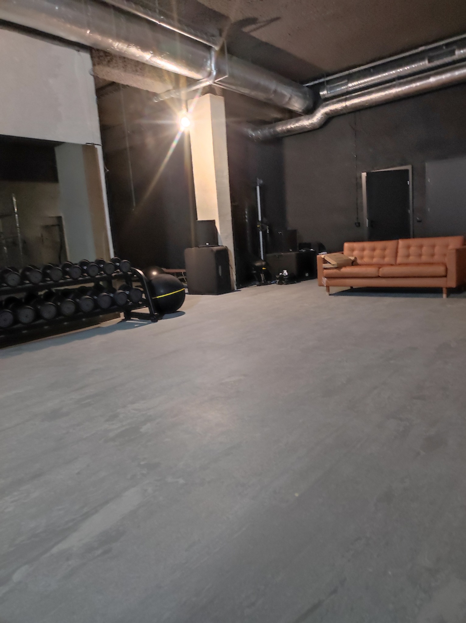 Przestronne, surowe wnętrze z betonową podłogą, widoczne rzędy hantli na stojaku, czarne głośniki i brązowa sofa w tle, oświetlone mocnym światłem lampy.