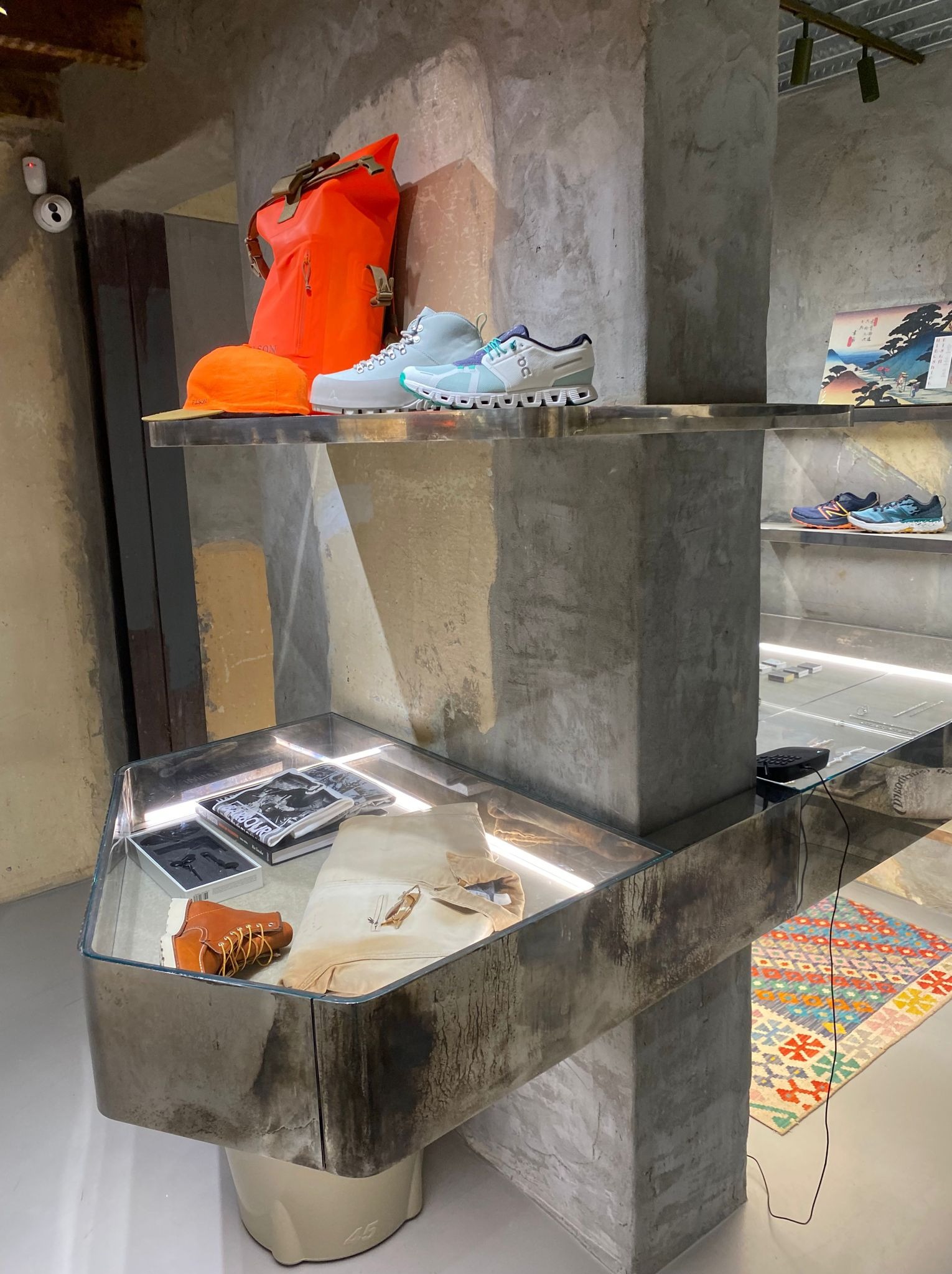 Ekspozycja w sklepie z odzieżą i obuwiem: pomarańczowy plecak, czapka i jasnoszare buty sportowe na półce z surowego betonu, poniżej witryna z brązowymi butami sznurowanymi, książkami i ubraniami...