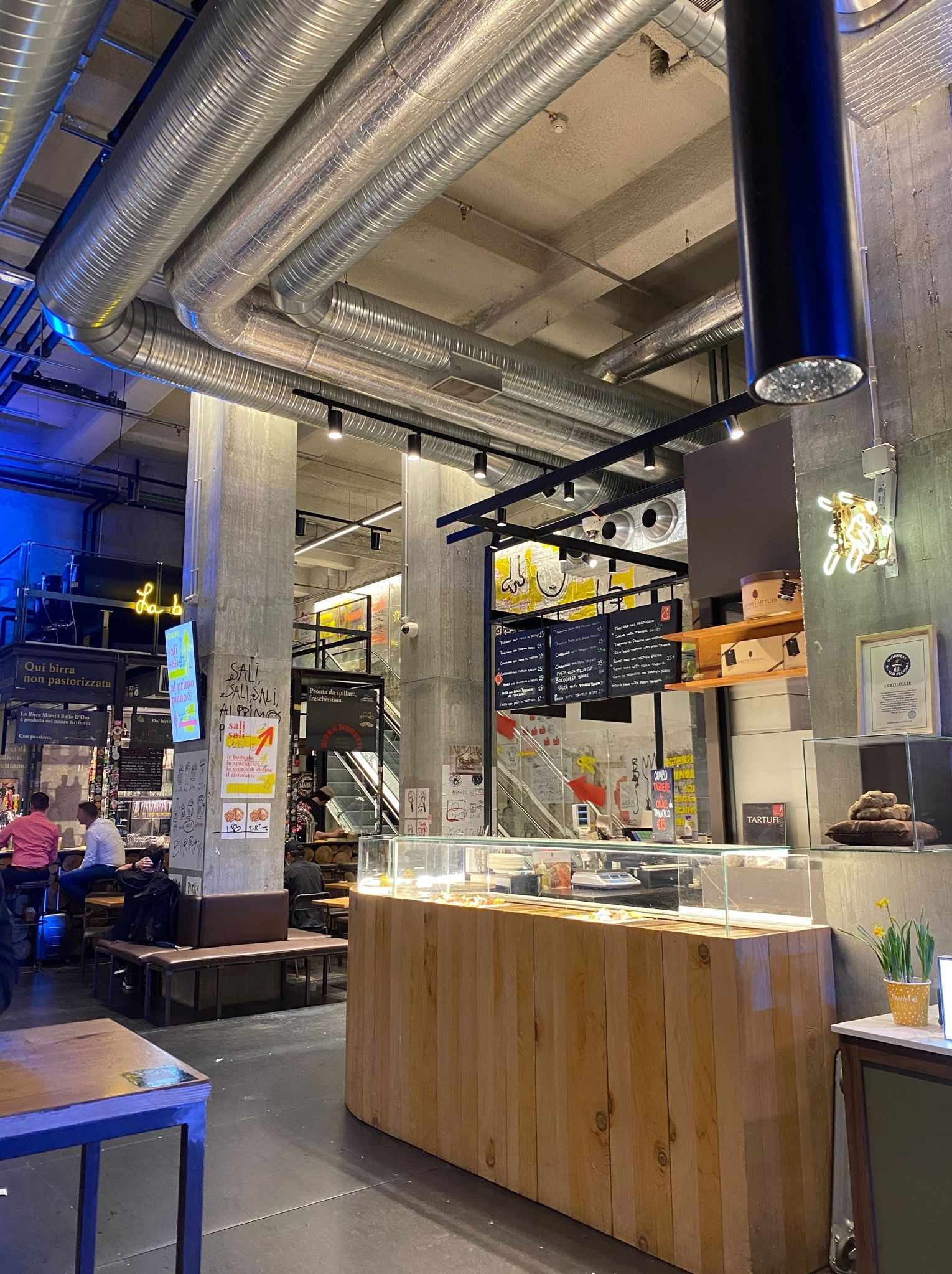 Wnętrze hali targowej z widocznymi instalacjami wentylacyjnymi pod sufitem, betonowymi kolumnami z napisami, oświetleniem punktowym, ladą chłodniczą i neonowym szyldem.