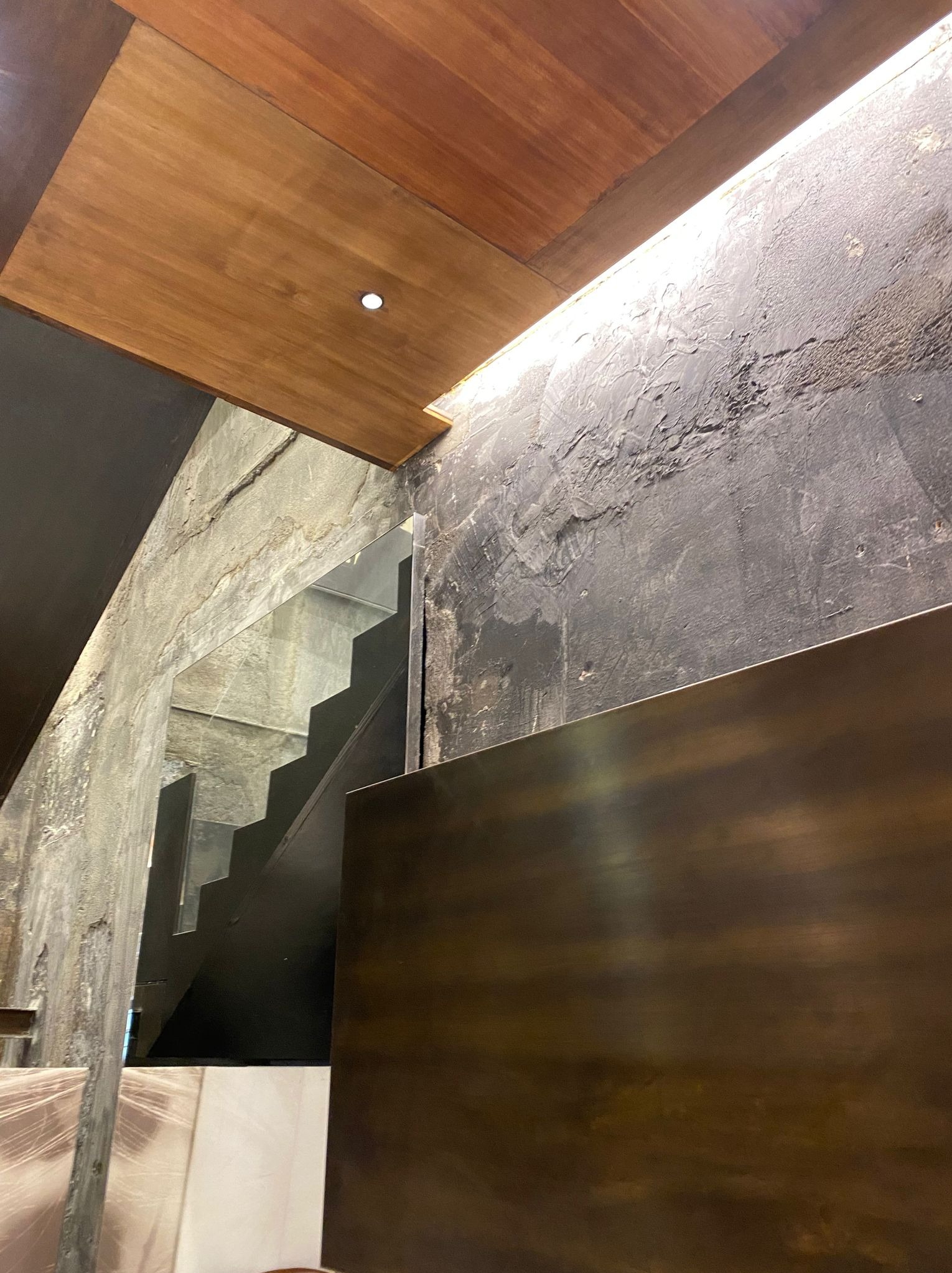 Surowe, teksturowane ściany w odcieniach szarości kontrastują z ciepłym drewnianym sufitem i elementami schodów z ciemnego metalu. Widoczne oświetlenie punktowe i fragment schodów z przeszkloną...