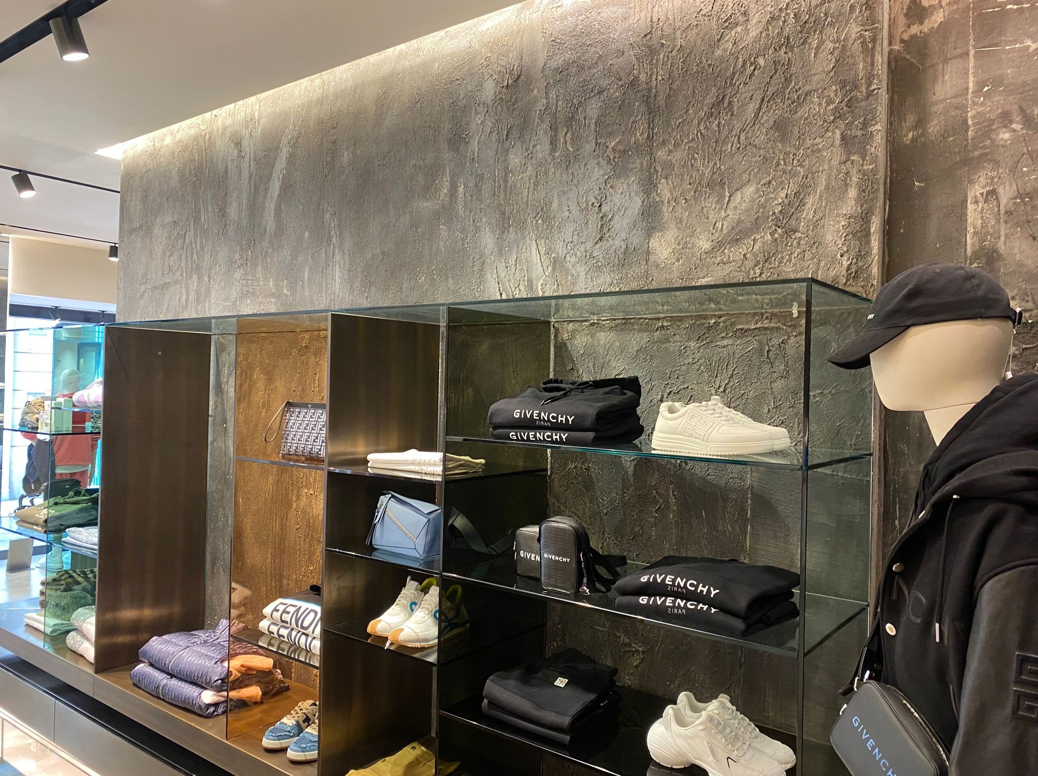 Wnętrze sklepu odzieżowego z półkami ekspozycyjnymi, na których znajdują się ubrania i buty marki Givenchy, tło stanowi ściana o surowej, betonowej fakturze, oświetlenie punktowe. Manekin w czapce...