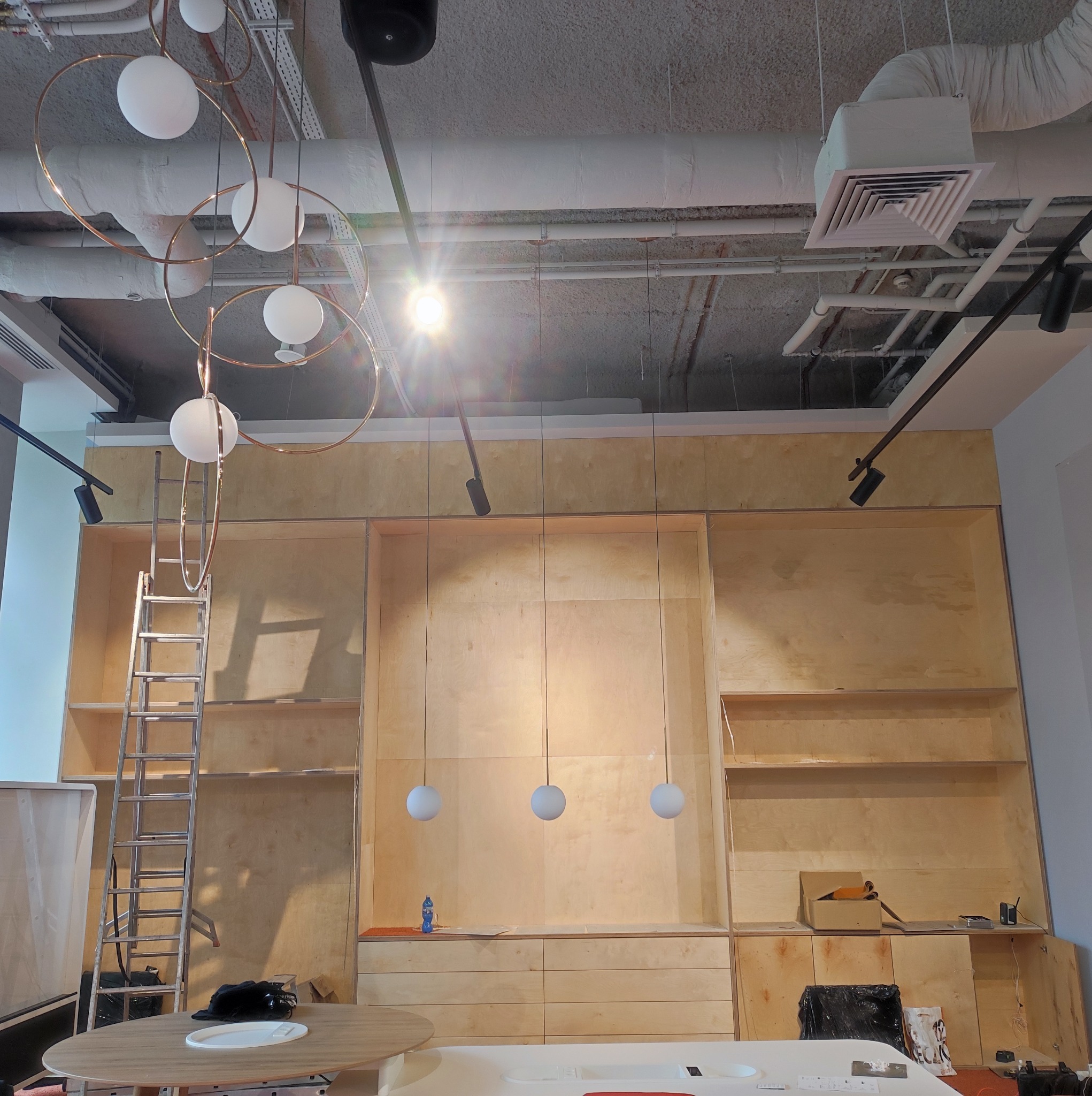 Wnętrze w trakcie wykańczania: surowe drewniane panele zabudowy ściennej z półkami, drabina oparta o ścianę, designerska lampa wisząca z okręgami i kulami, widoczny sufit z instalacjami.