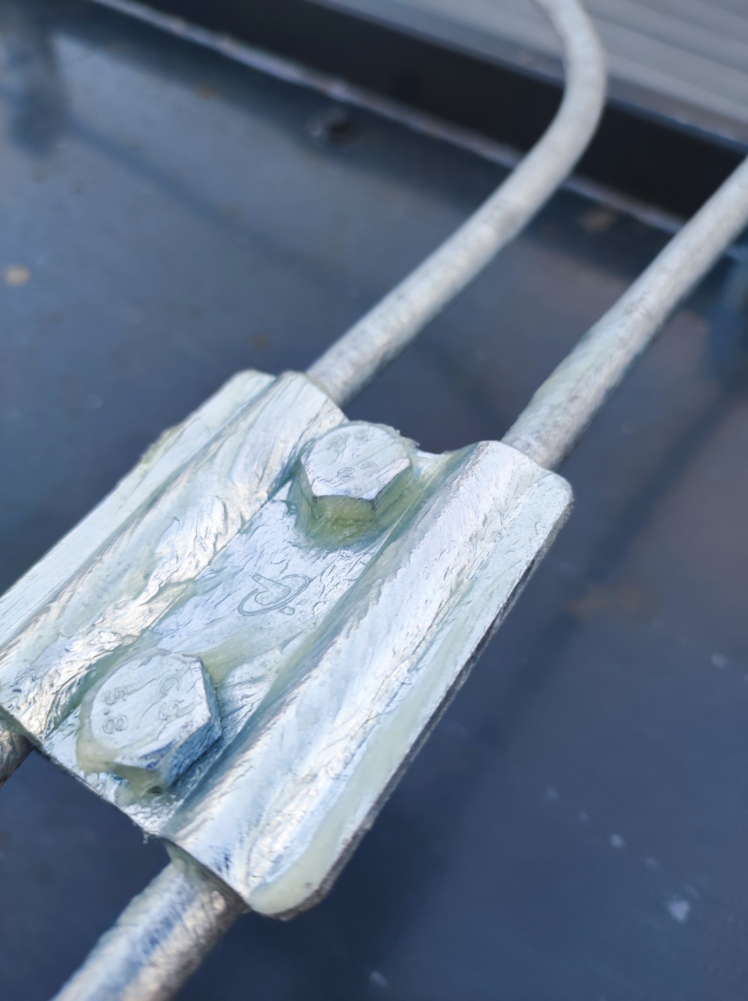 Metalowy zacisk mocujący dwa przewody, zabezpieczony śrubami, widoczne detale konstrukcyjne i mocowania.