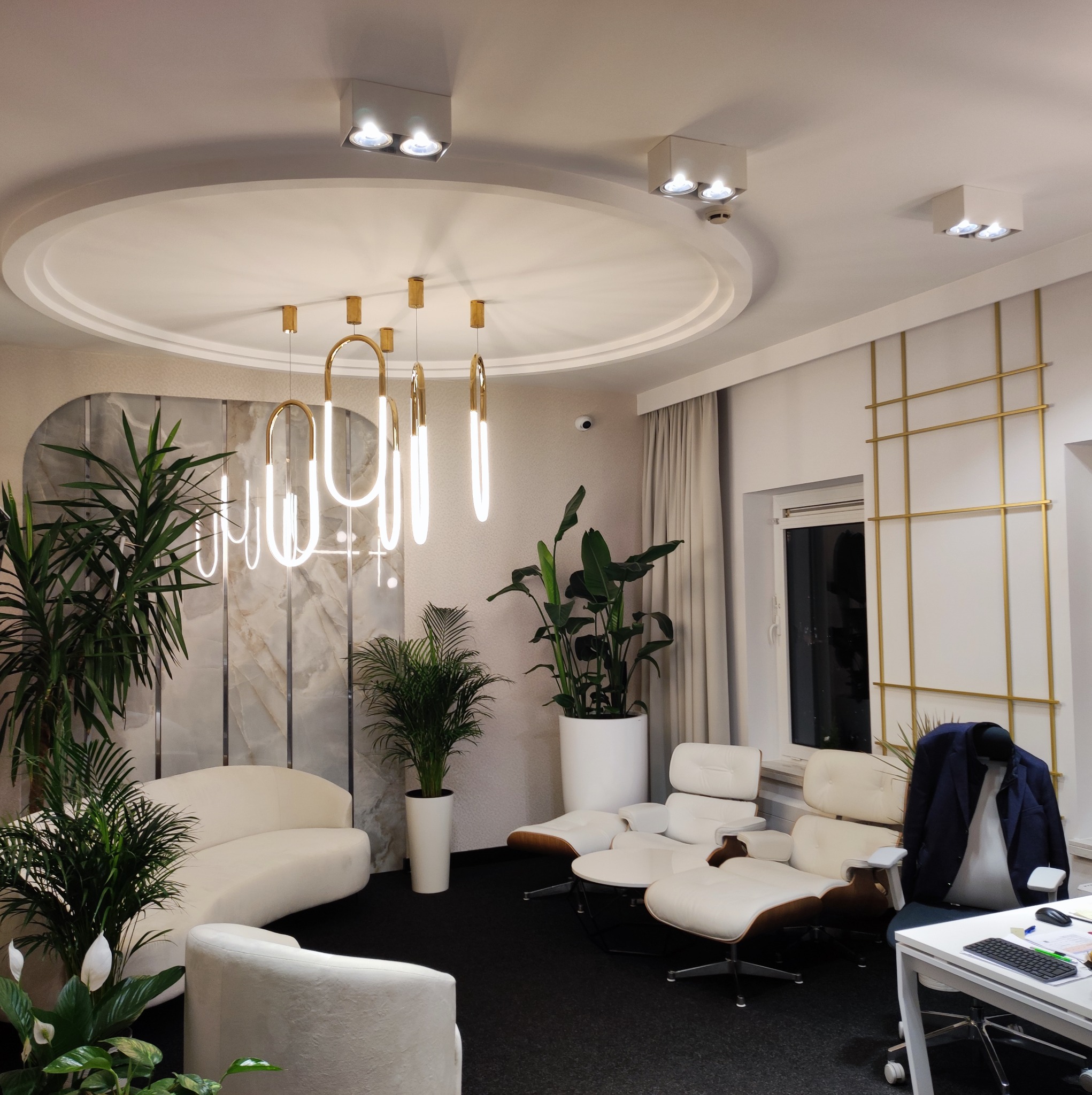 Eleganckie wnętrze biurowe z nowoczesnym oświetleniem wiszącym, białymi meblami wypoczynkowymi, roślinami doniczkowymi i białymi ścianami z geometrycznymi złotymi akcentami.