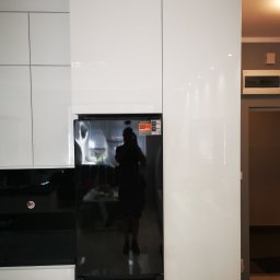 MK FURNITURE Michał Kalinowski - Biała zabudowa kuchenna z błyszczącymi frontami, wkomponowana czarna lodówka Indesit z naklejkami informacyjnymi, odbicie osoby w lodówce.