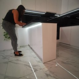 MK FURNITURE Michał Kalinowski - Kobieta w czarnej koszulce i szarych dżinsach pracuje przy białej, nowoczesnej wyspie kuchennej z czarnym blatem, oświetloną od spodu, w minimalistycznej kuchni z białymi szafkami i marmurową podłogą.