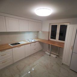 MK FURNITURE Michał Kalinowski - Jasna kuchnia na wymiar z drewnianym blatem, białymi frontami szafek z frezowanymi detalami i złotymi uchwytami, marmurowa podłoga, widoczny fragment grzejnika pod blatem przy oknie.