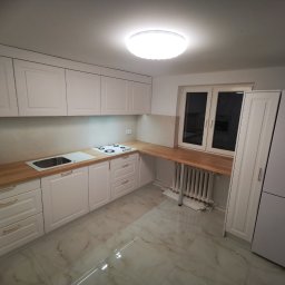 MK FURNITURE Michał Kalinowski - Jasna kuchnia z drewnianym blatem, białymi frontami szafek z frezowanymi ramkami, złotymi uchwytami, płytą grzewczą i zlewem, widok na grzejnik pod blatem przy oknie.