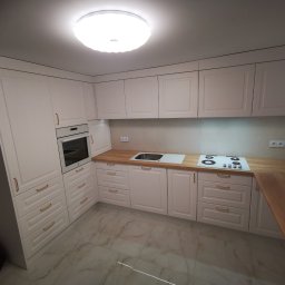 MK FURNITURE Michał Kalinowski - Jasna kuchnia z drewnianymi blatami, fronty szafek z frezowanymi detalami, złote uchwyty, marmurowe płytki podłogowe, oświetlenie sufitowe LED.