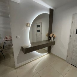 MK FURNITURE Michał Kalinowski - Nowoczesna aranżacja przedpokoju z okrągłym lustrem podświetlanym LED, konsolą z szufladami i dekoracyjnym oświetleniem ściennym. W tle widoczny pokój z krzesełkiem do karmienia.