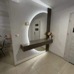 Nowoczesna aranżacja przedpokoju z okrągłym lustrem podświetlanym LED, konsolą z szufladami i dekoracyjnym oświetleniem ściennym. W tle widoczny pokój z krzesełkiem do karmienia.