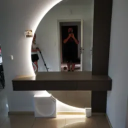 Nowoczesna toaletka z podświetlanym lustrem w kształcie półksiężyca, odbicie osoby robiącej zdjęcie telefonem, w tle osoba odkurzająca.