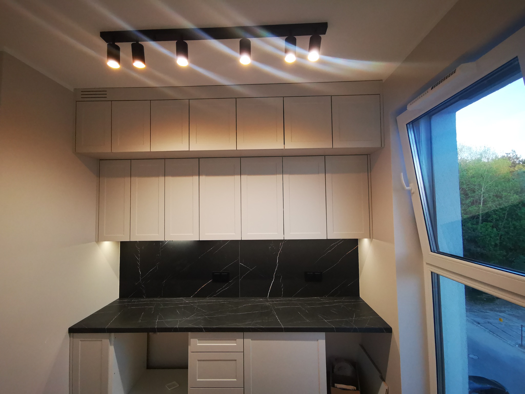 Fragment kuchni z białymi szafkami, blatem imitującym czarny marmur i oświetleniem punktowym, widok z boku na otwarte okno z widokiem na zewnątrz.
