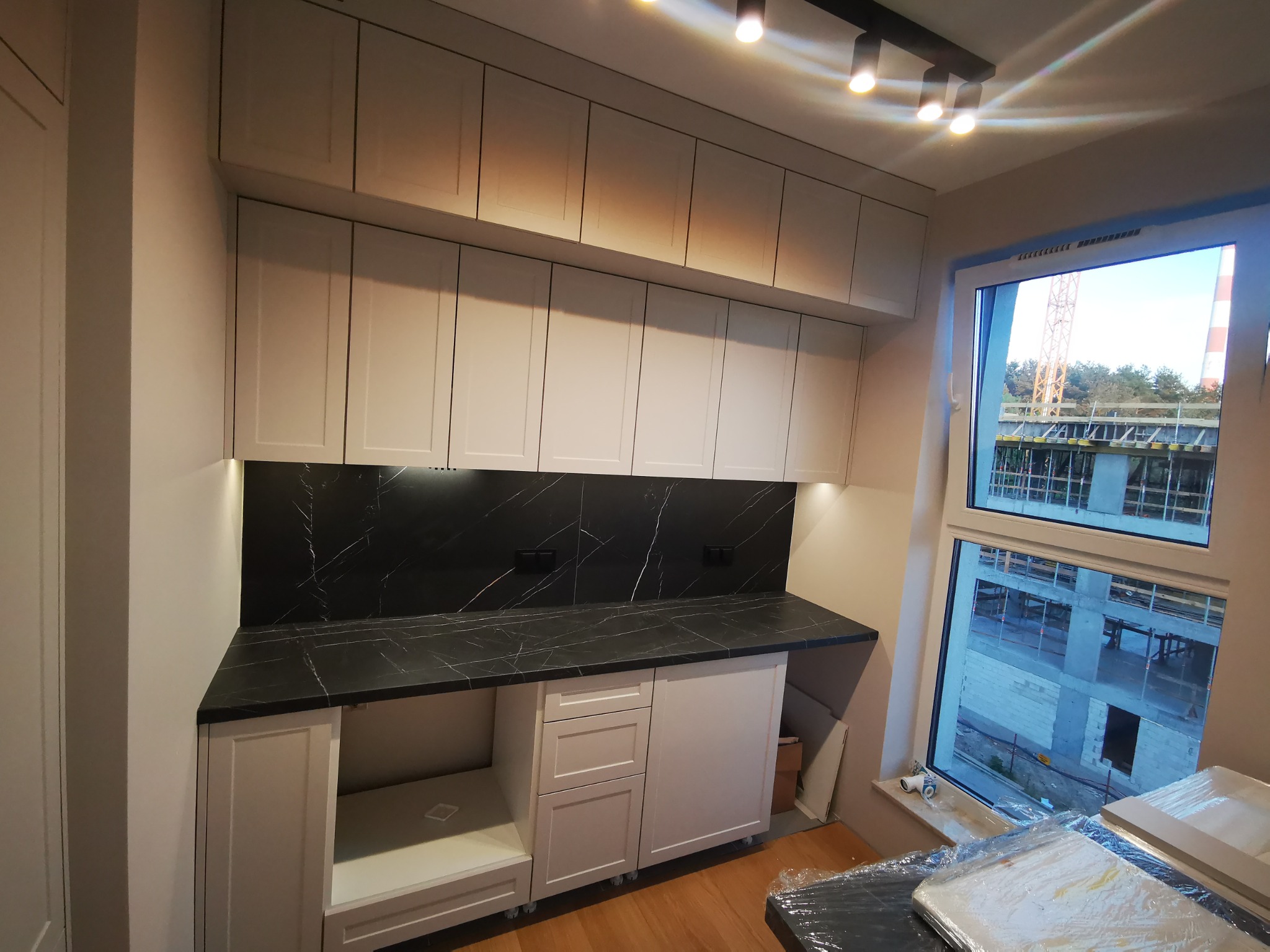 Jasna zabudowa kuchenna z czarnym blatem imitującym marmur, górne szafki podwieszane pod sufitem, widok na budowę za oknem.