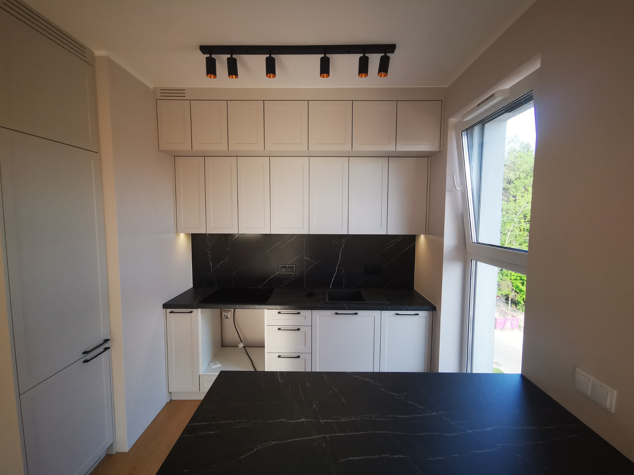 Jasna kuchnia z czarnym blatem i backsplashiem imitującym marmur, oświetlona czarnymi reflektorami sufitowymi, z widocznym fragmentem wyspy kuchennej i otwartym oknem.