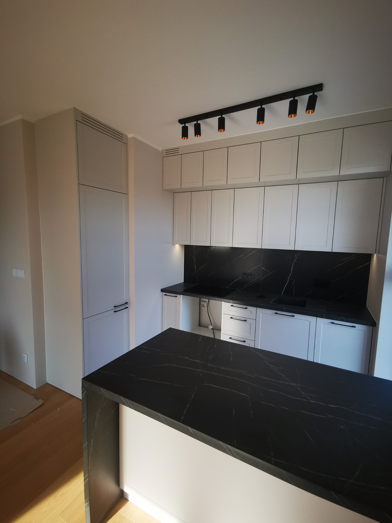 Nowoczesna kuchnia z białymi szafkami, czarnym blatem z marmurowym wzorem i oświetleniem punktowym na suficie.