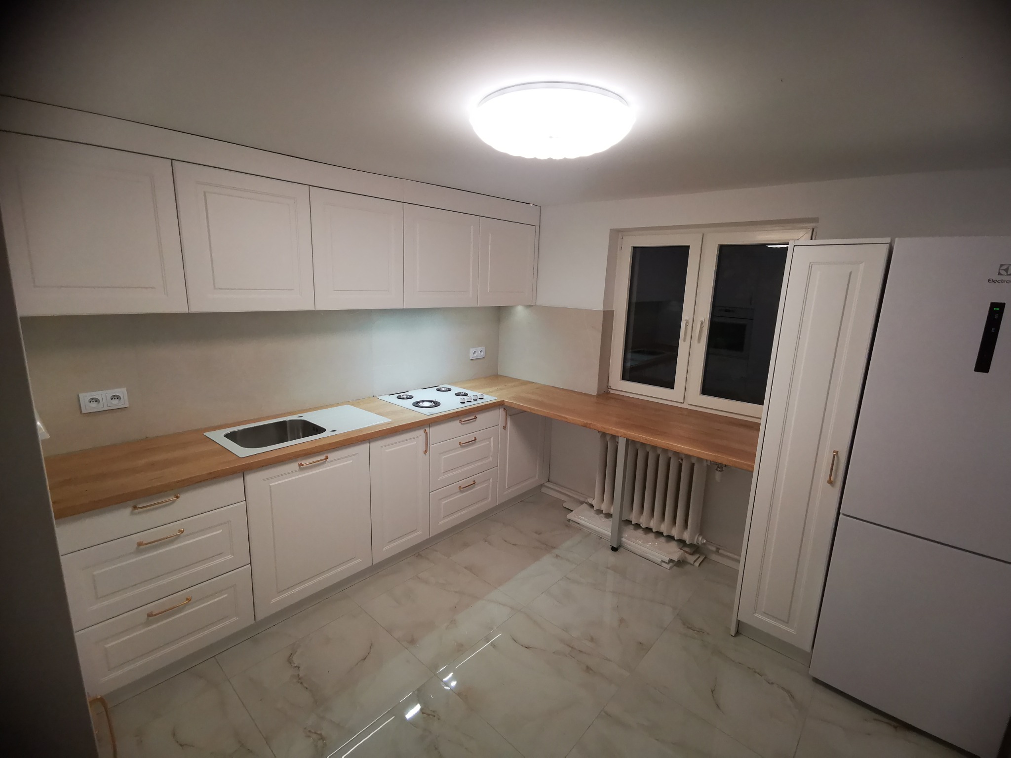 Jasna kuchnia na wymiar z drewnianym blatem, białymi frontami szafek z frezowanymi detalami i złotymi uchwytami, marmurowa podłoga, widoczny fragment grzejnika pod blatem przy oknie.