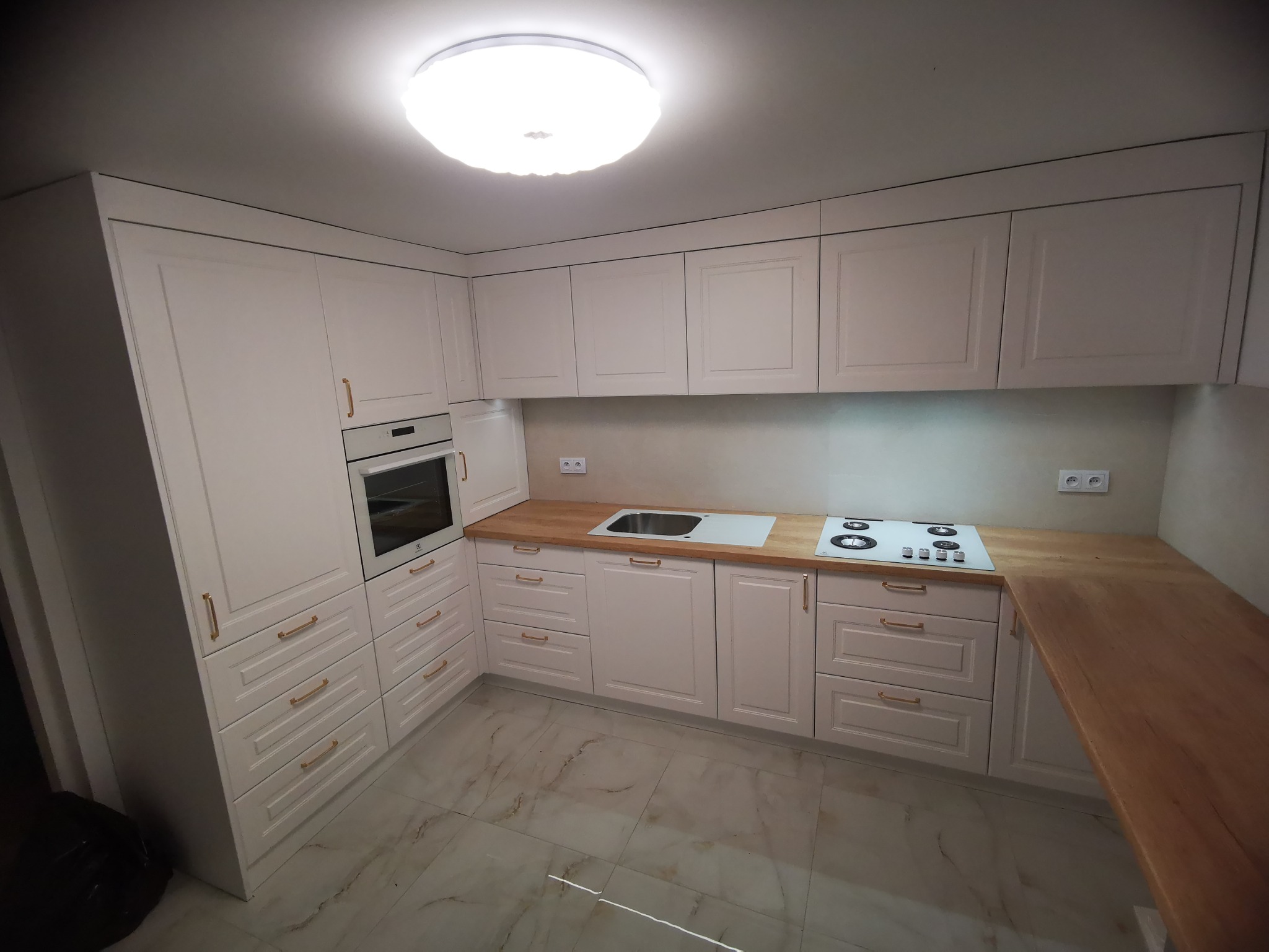 Jasna kuchnia z drewnianymi blatami, fronty szafek z frezowanymi detalami, złote uchwyty, marmurowe płytki podłogowe, oświetlenie sufitowe LED.