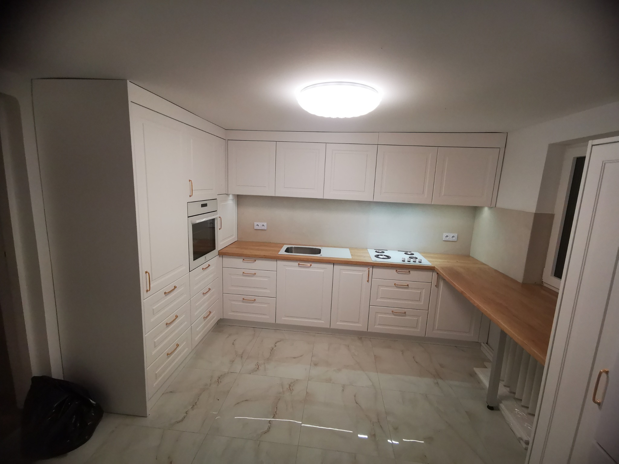 Jasna kuchnia z drewnianymi blatami, frezowane fronty szafek, złote uchwyty, marmurowe płytki podłogowe, okap zintegrowany z szafką, piekarnik w zabudowie.