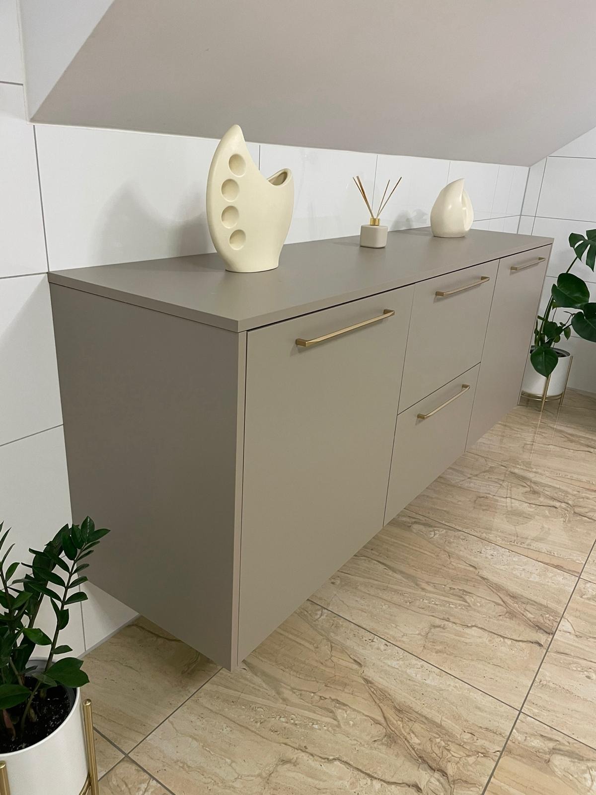 Beżowa szafka wisząca z złotymi uchwytami na tle białych płytek i marmurowej podłogi, udekorowana białymi wazonami i roślinami doniczkowymi.
