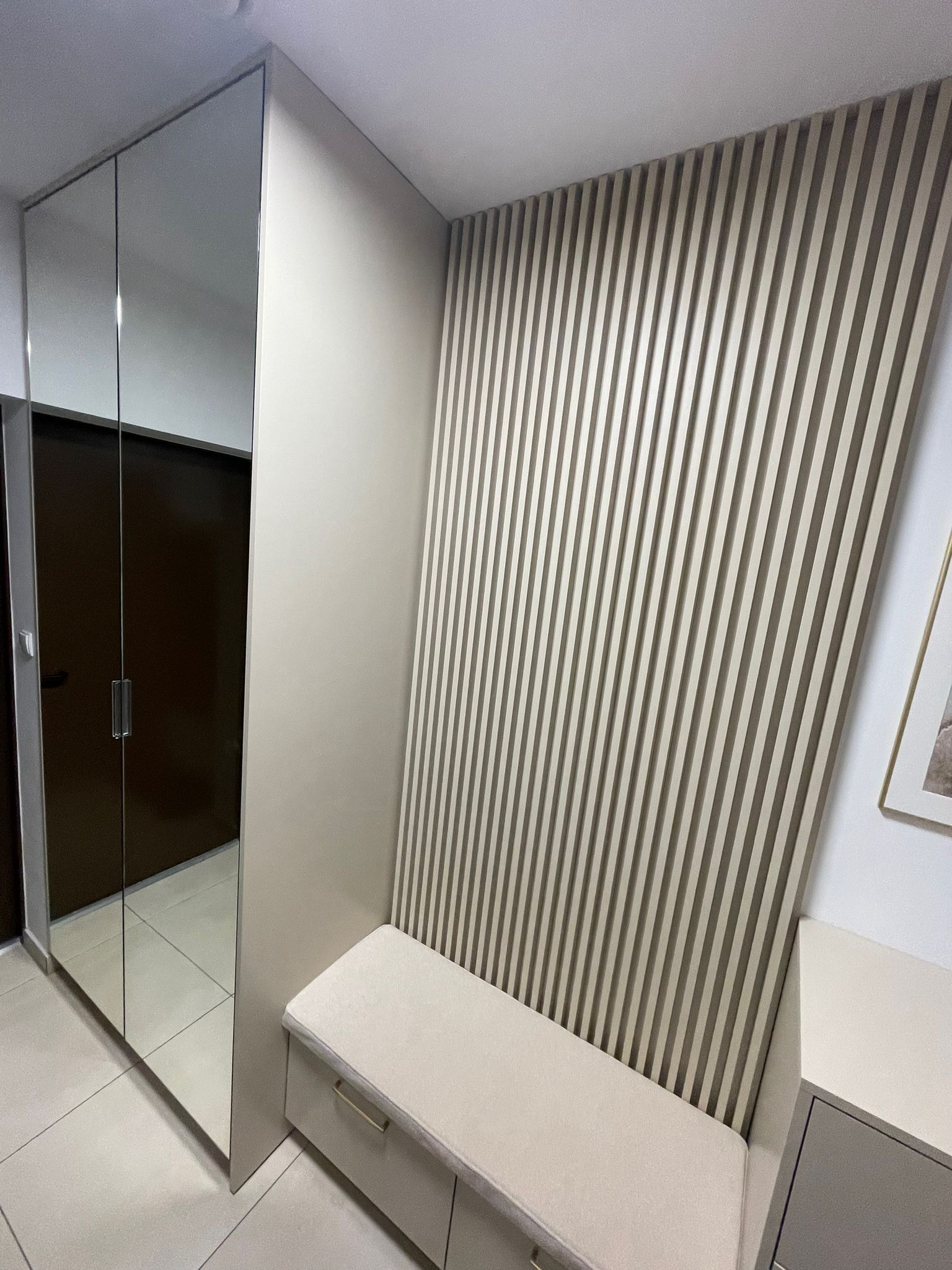 Nowoczesna szafa wnękowa z lustrzanymi drzwiami przesuwnymi, połączona z panelem dekoracyjnym z pionowych listew w odcieniach beżu i siedziskiem z pojemnikiem, tworząca spójną aranżację przedpokoju.