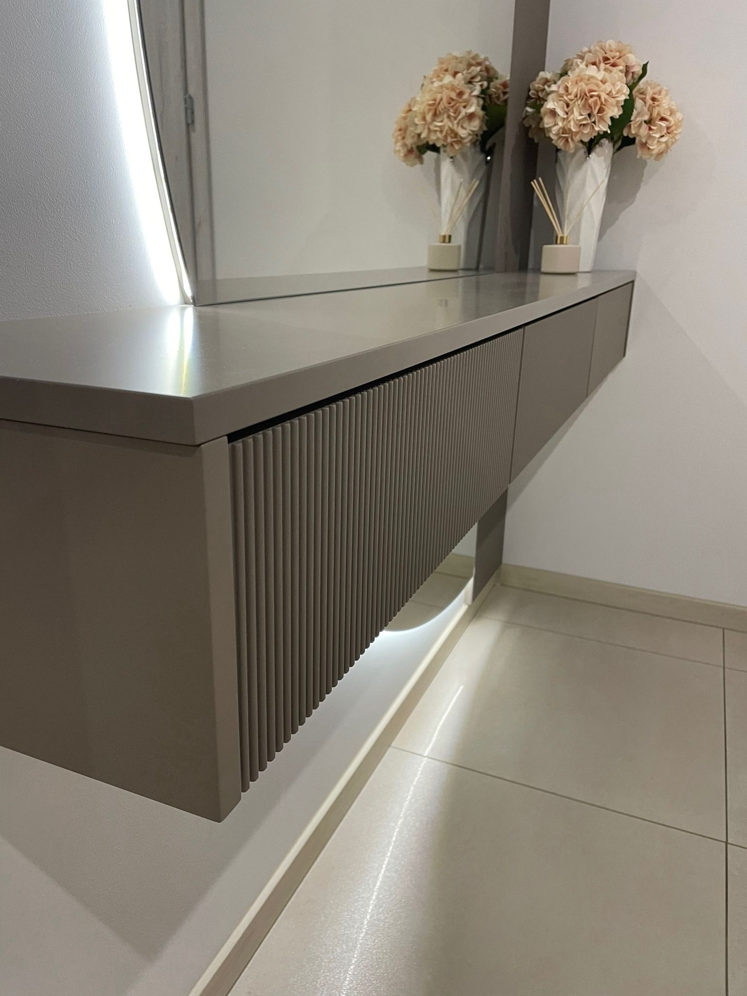 Szara, wisząca konsola z ryflowanym frontem, podświetlona od spodu, odbijająca się w lustrze z wbudowanym oświetleniem LED, na której stoją dekoracyjne kwiaty w wazonach.