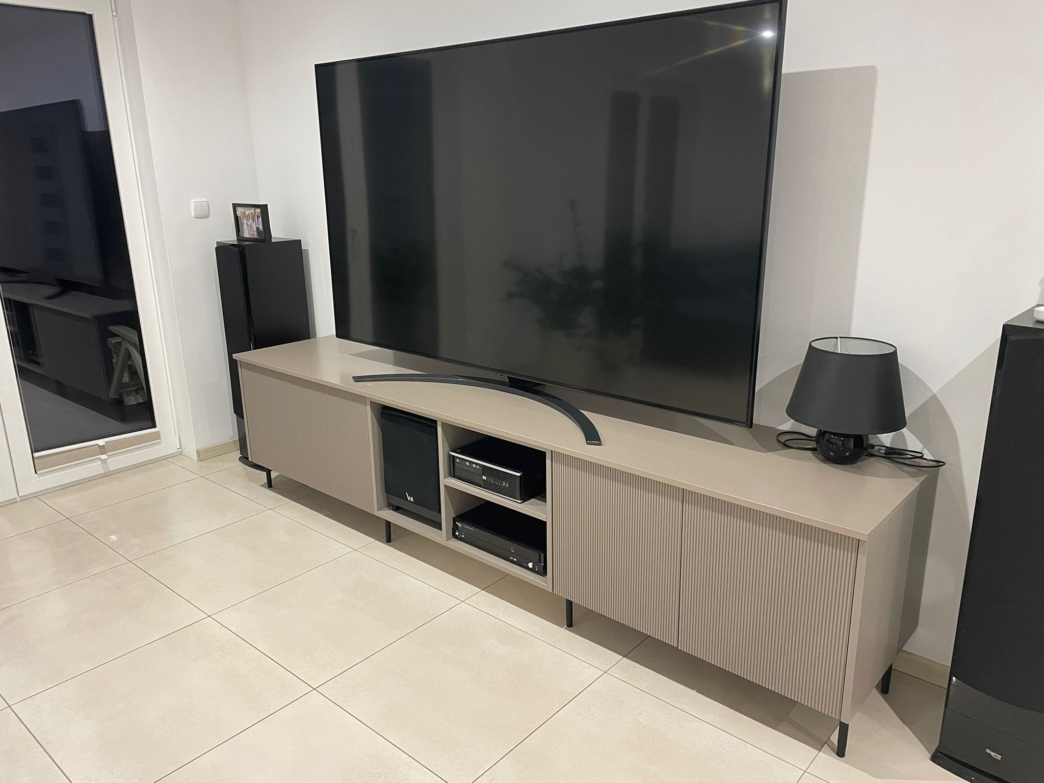 Beżowa szafka RTV z ryflowanymi drzwiczkami, na cienkich nóżkach, z czarnym telewizorem, głośnikami i lampką, na jasnych płytkach podłogowych.